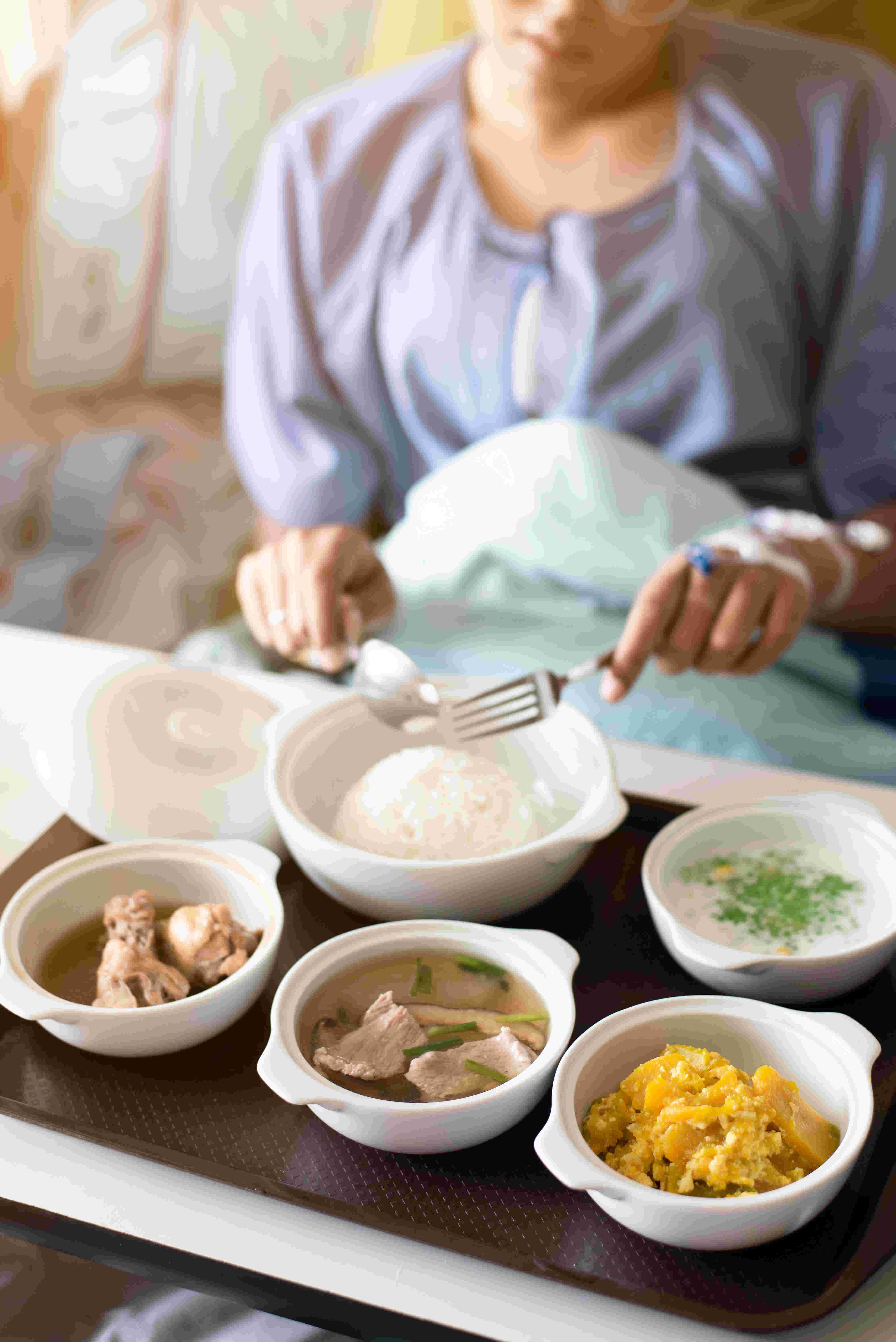 Une femme mange un plateau-repas : cinq plats en porcelaine blanche, certains avec couvercle, contenant des pâtes, du riz, de la viande...