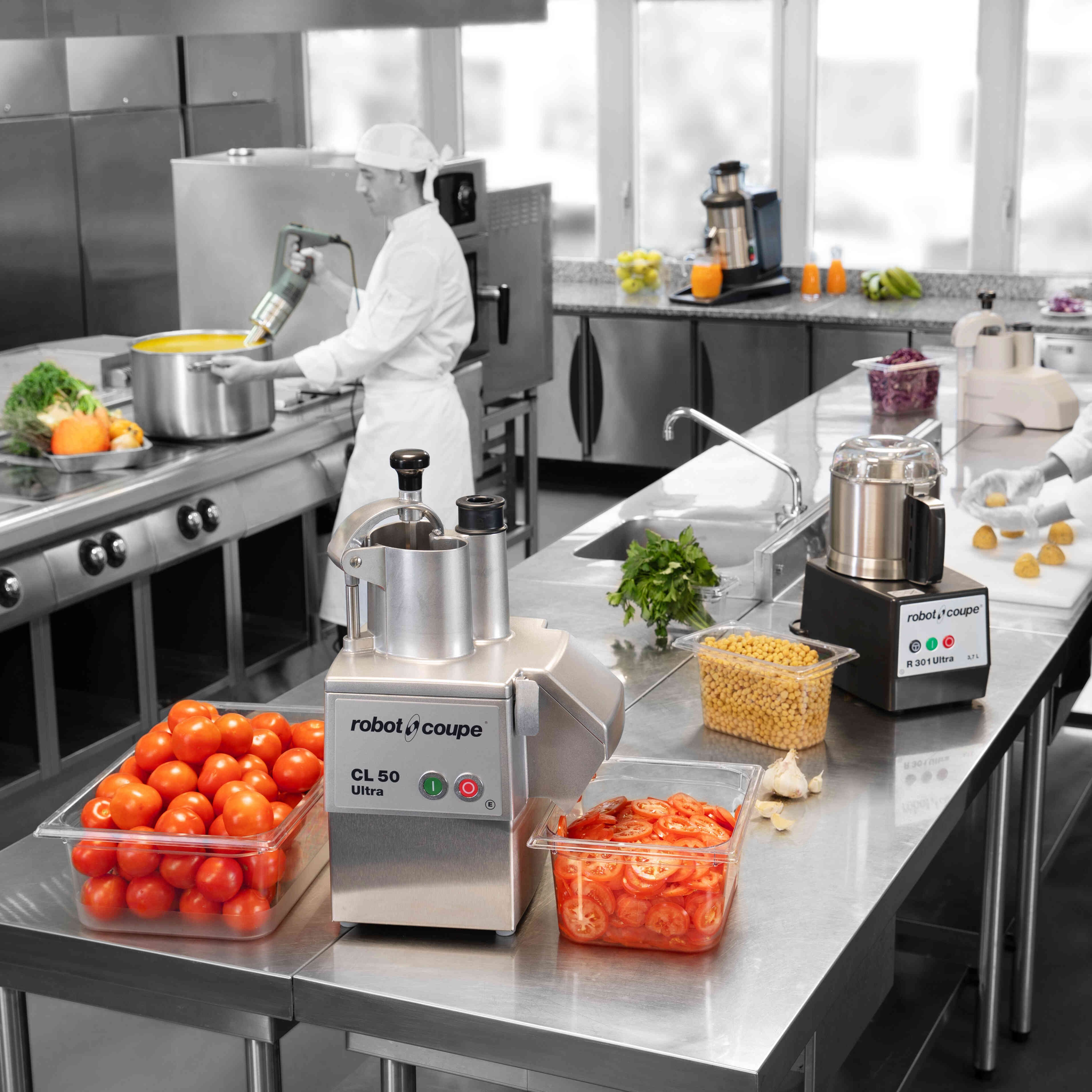 Vue générale d'une cuisine professionnelle. Sur le plan de travail, des tomates tranchées et non-tranchées et deux coupes-légumes électriques.