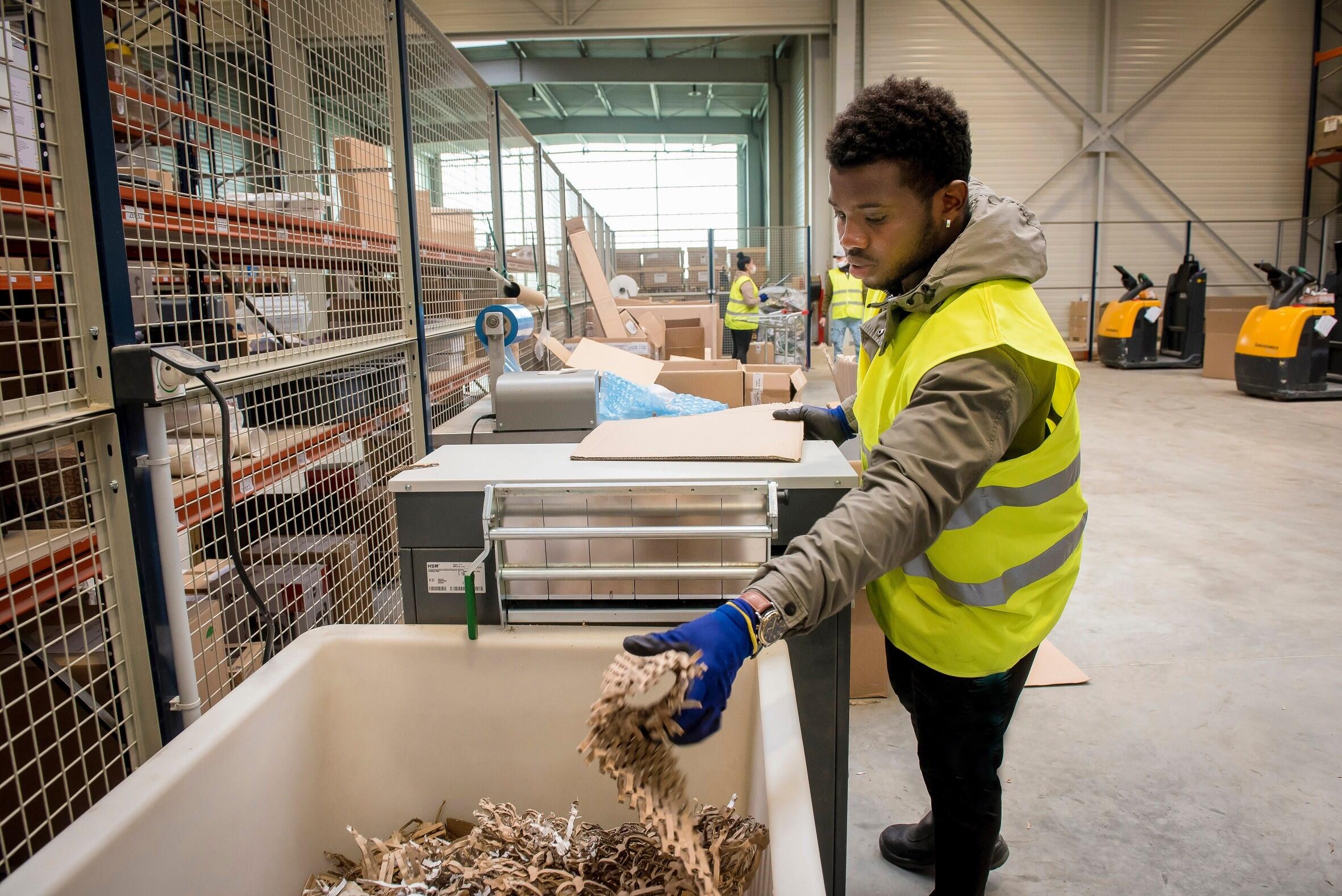 Homme vêtu d'une veste jaune de sécurité travaillant dans un entrepôt : il prend des chutes de carton pour protéger les produits lors du transport.