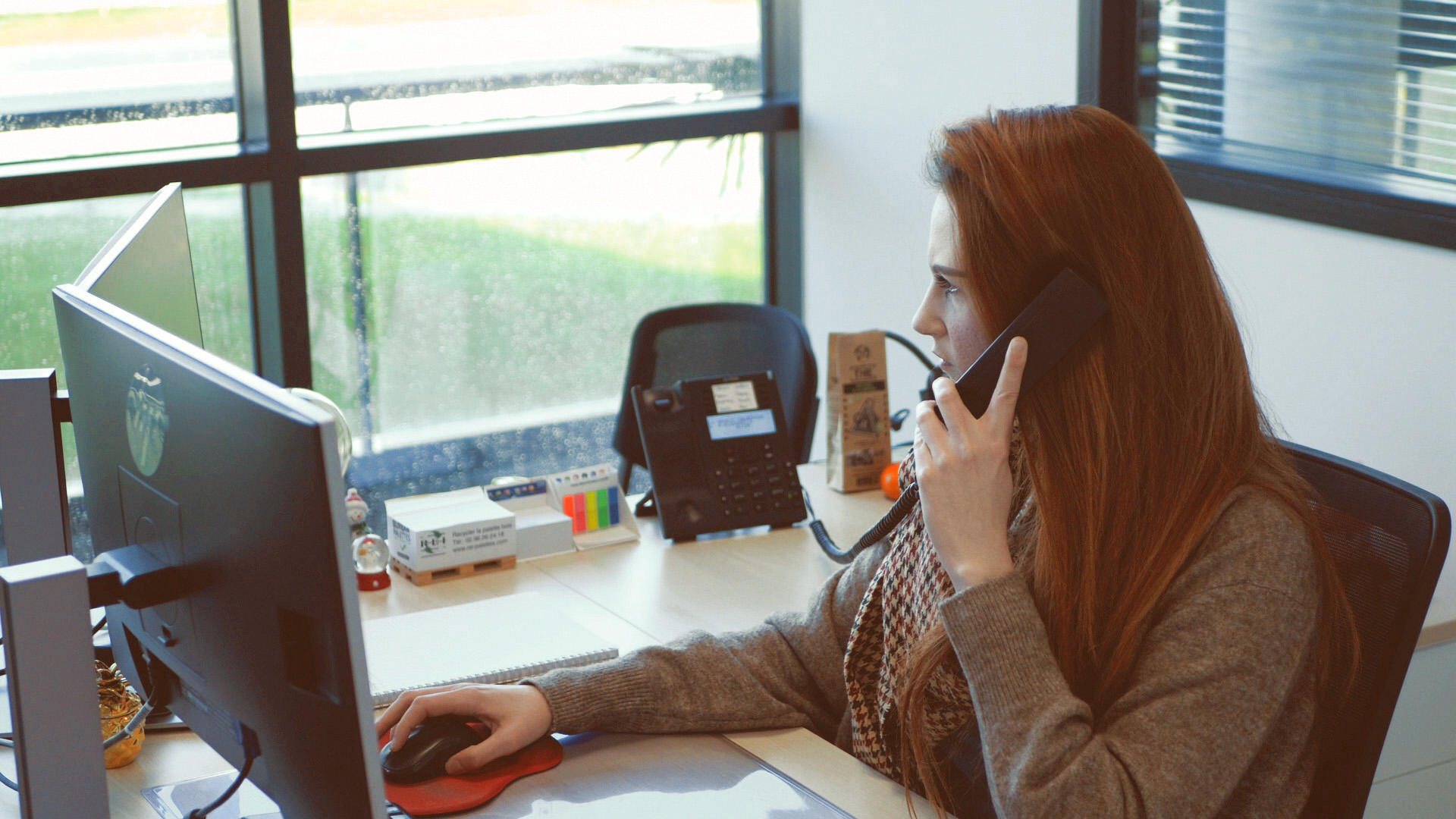Femme brune, la trentaine, assise à son bureau face à son écran d'ordinateur et parlant au téléphone.