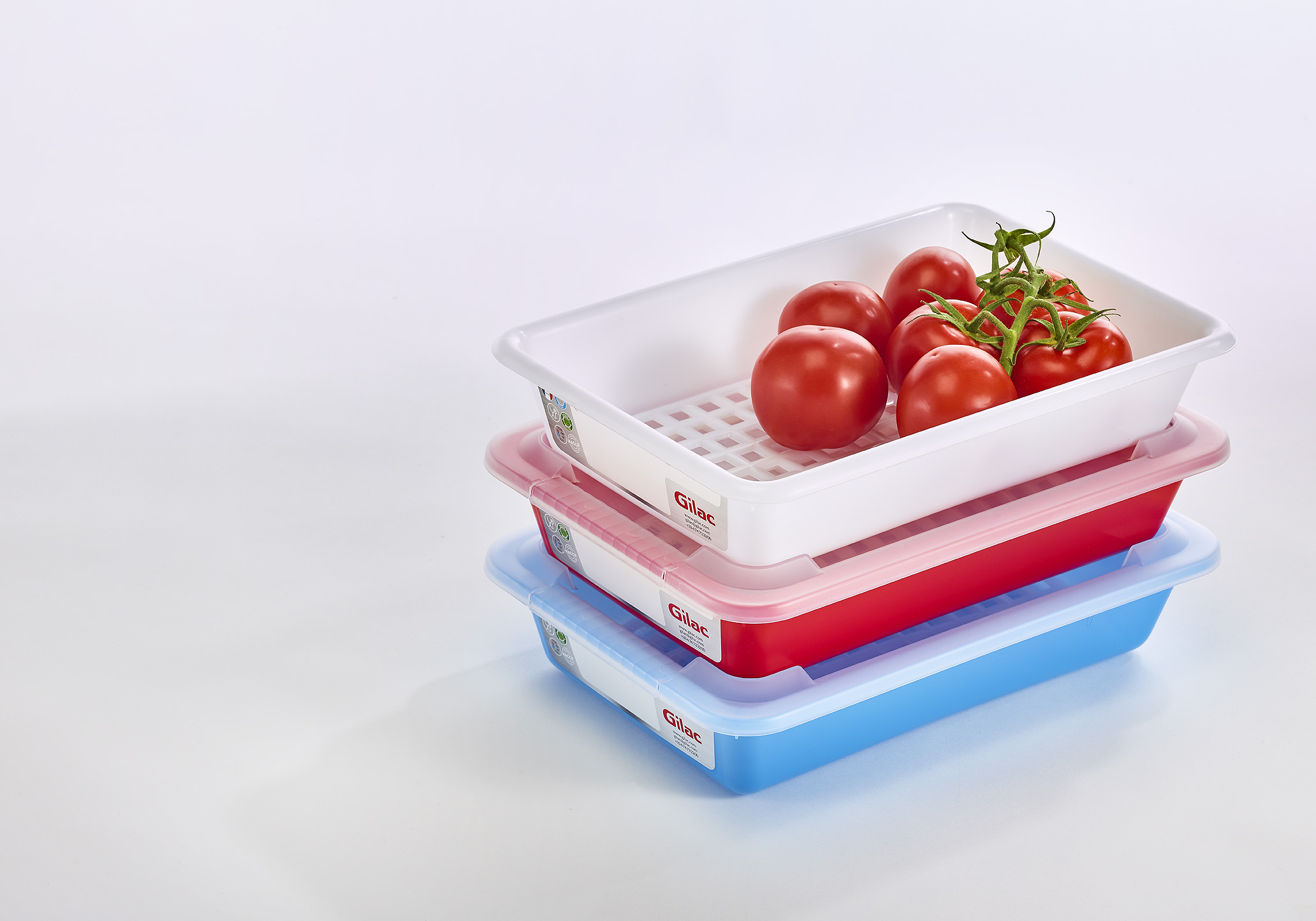 Trois bacs de stockages bleus, blancs et rouges de la marque Gilac contenant des tomates et au fond percé de trous.