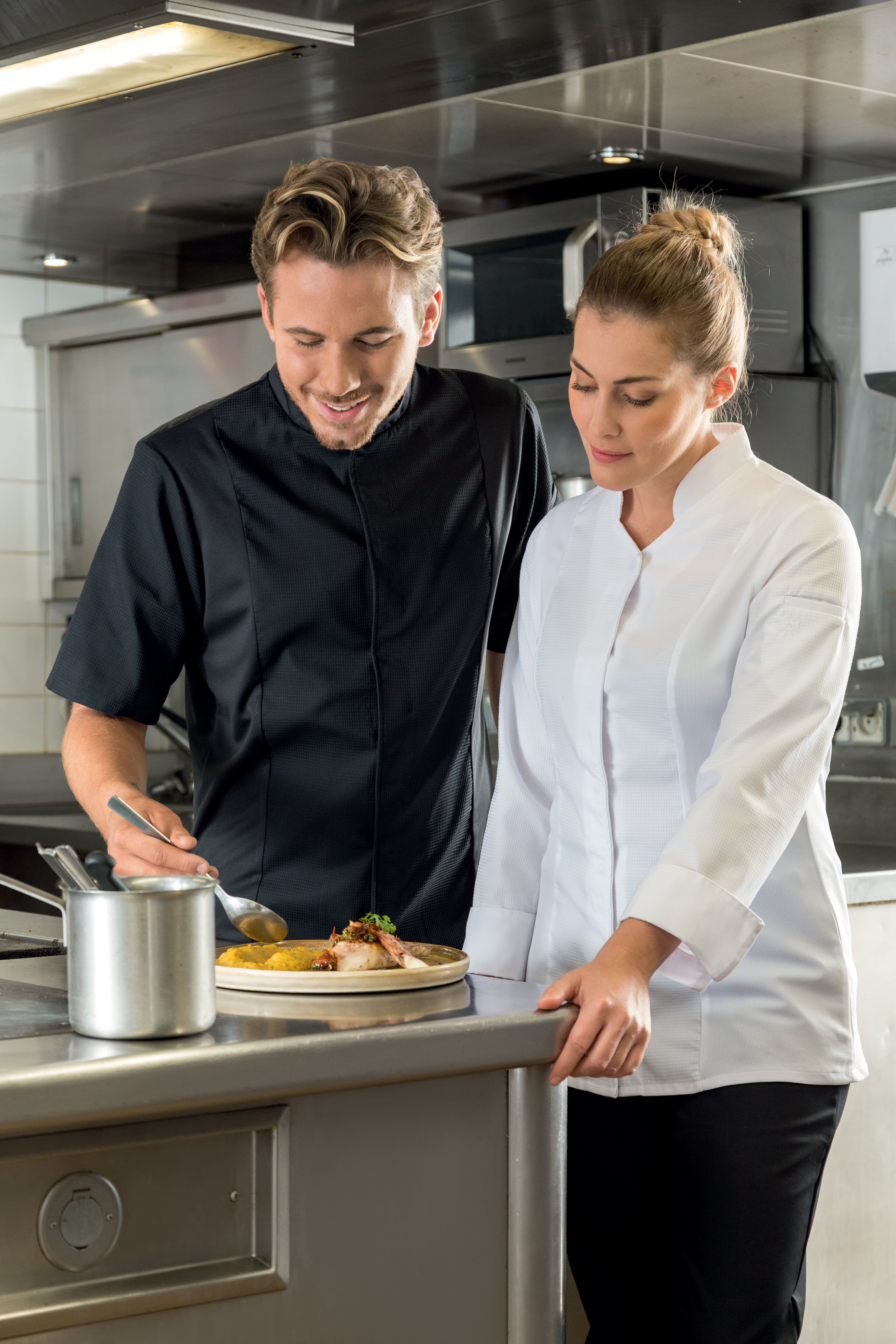 Un homme et une femme dans une cuisine, vêtus de veste et de pantalon de cuisine ; noirs pour lui, blancs pour elle ; de la marque Robur.