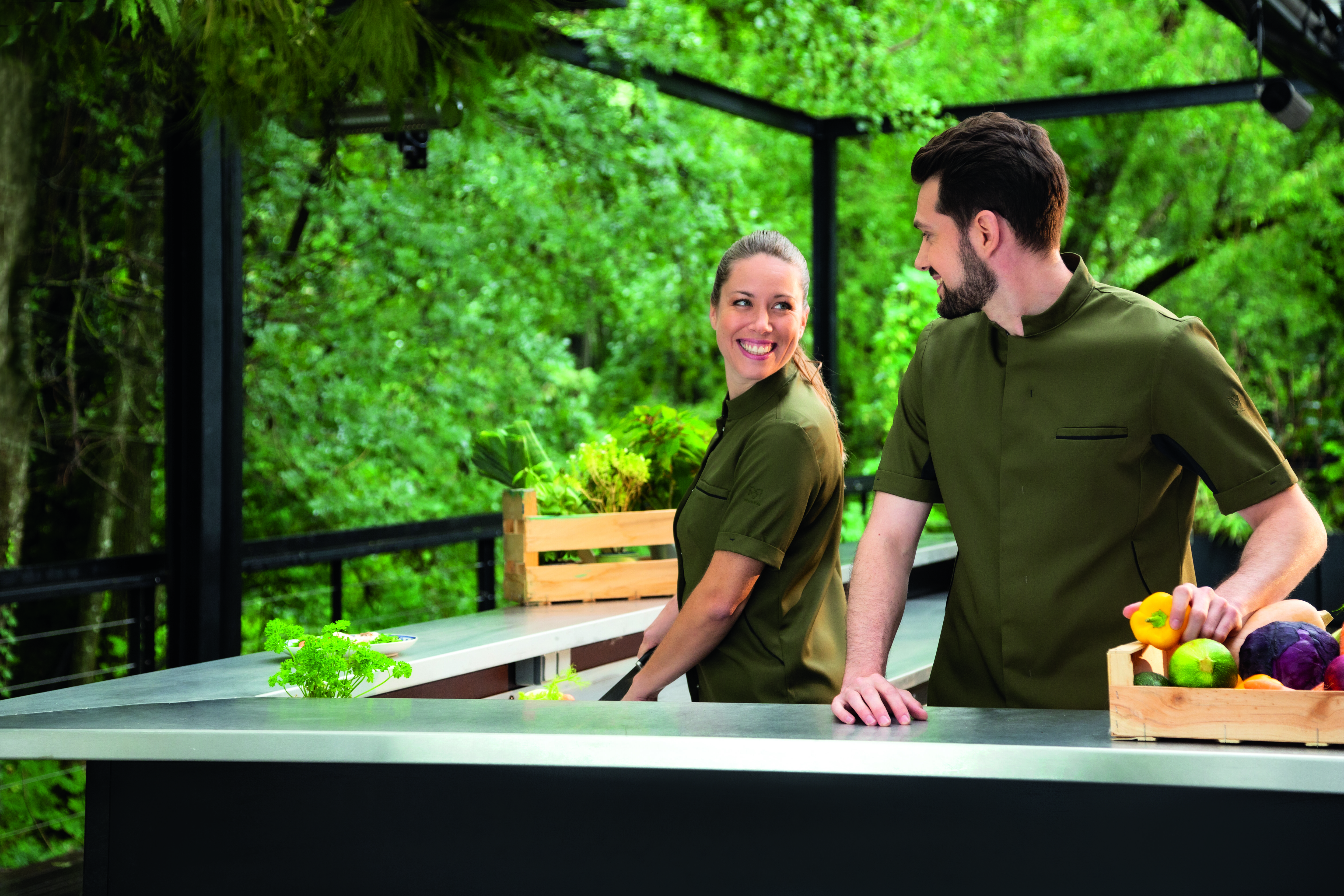 Deux serveurs dans un bar extérieurs, un homme et une femme. Ils portent chacun une chemisette vert kaki de la marque Robur.