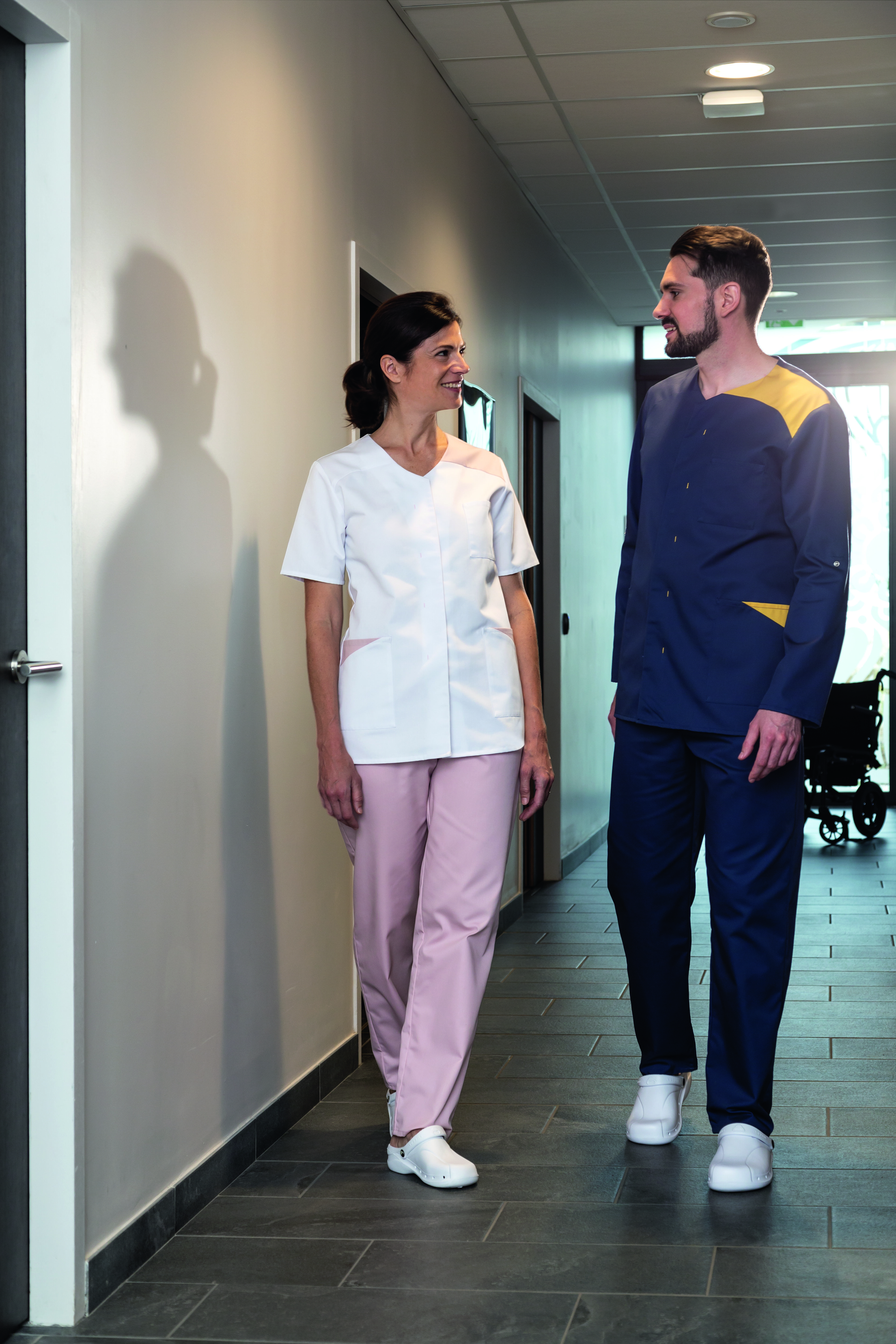 Un homme et une femme portent des vêtements de la marque Robur : une veste et un pantalon gris pour lui ; une veste et un pantalon rose pour elle.