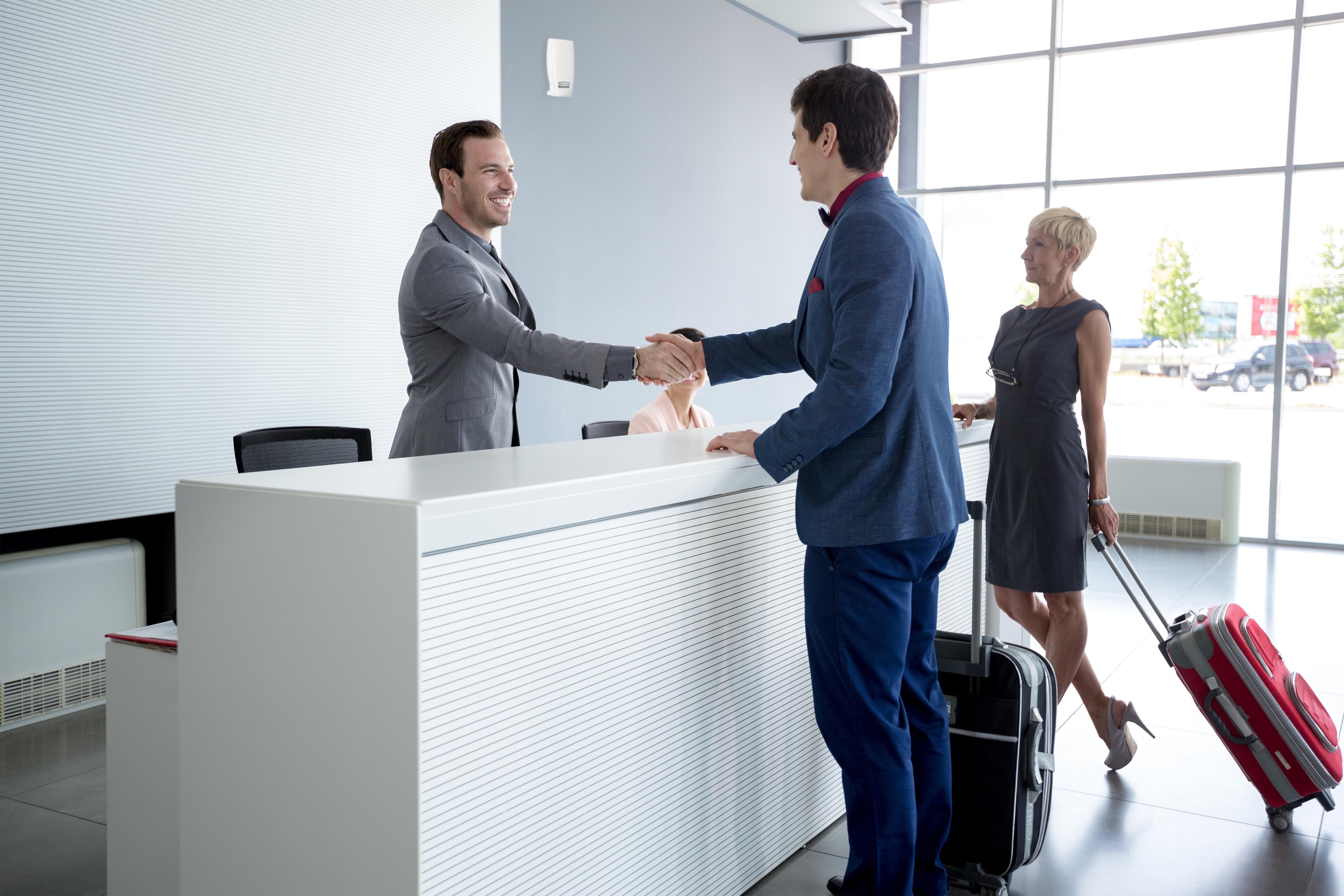 À un desk d'accueil, un homme serre la main d'une autre ; une femme s'approche avec une valise.