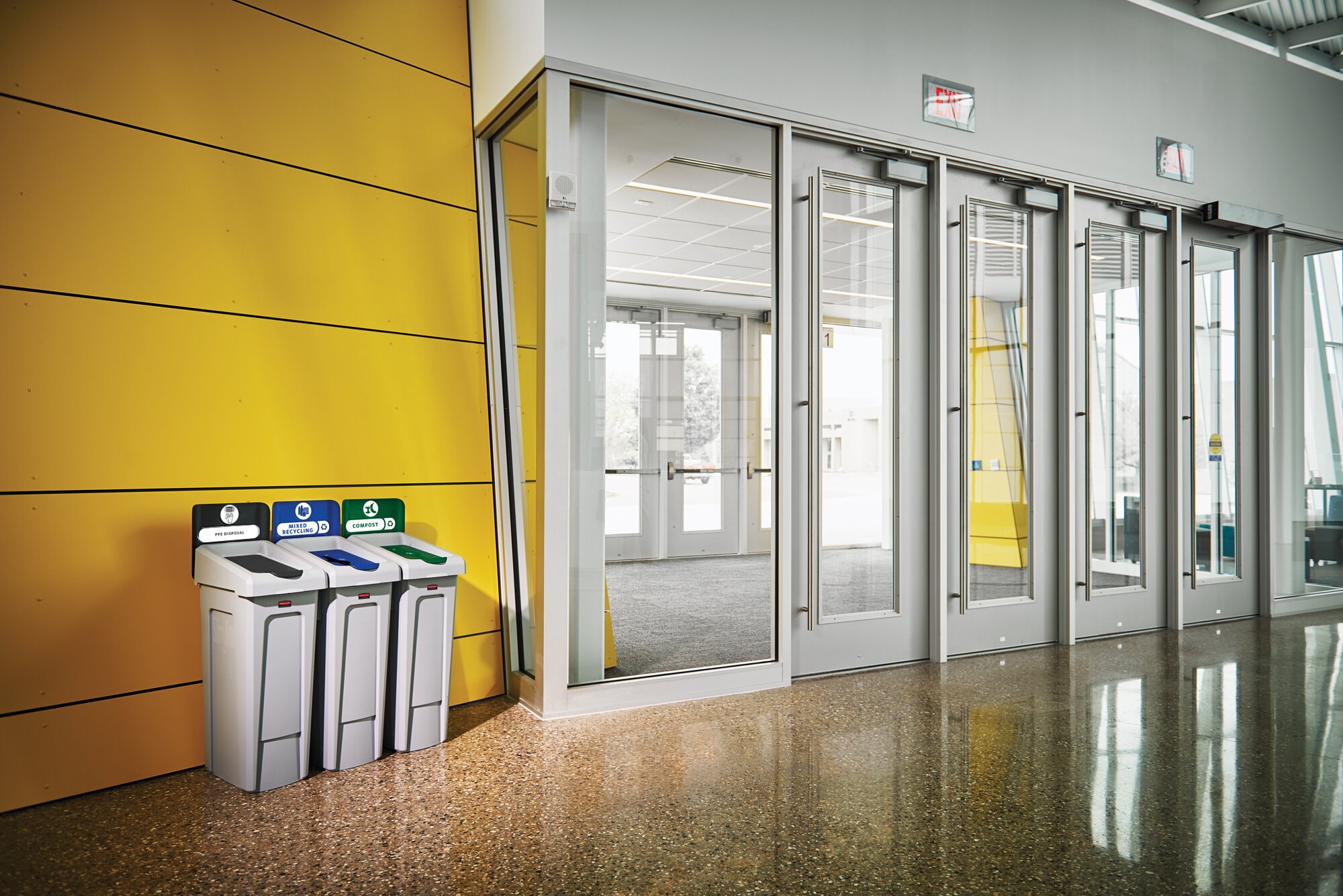 Dans un hall d'immeuble : trois bacs de tri à gauche de portes automatiques.