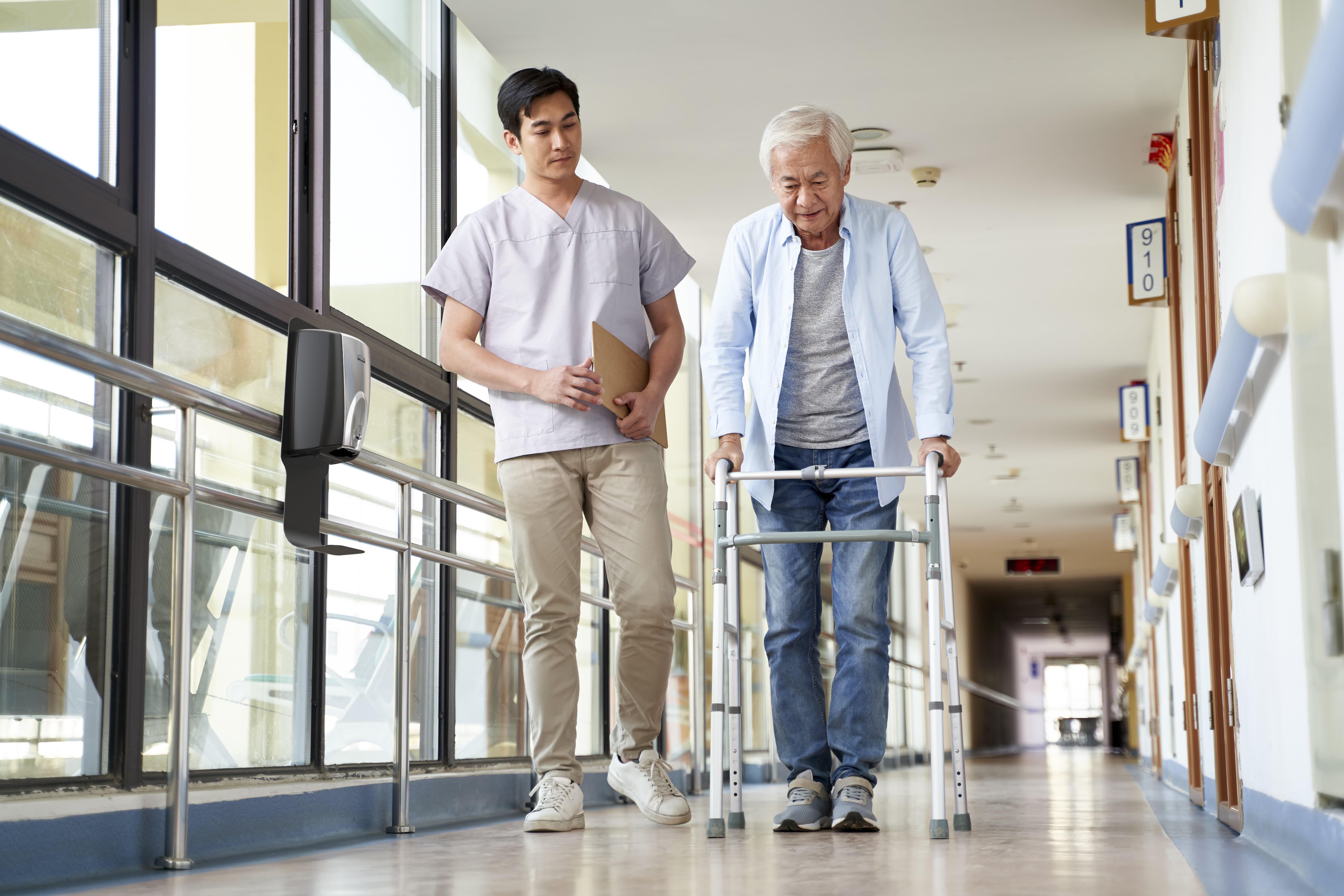 Dans le couloir d'un hôpital, un homme âgé marche avec un cadre. Un soignant l'accompagne. 